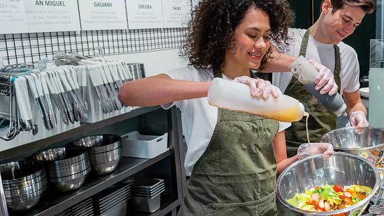 Two food service employees, wearing aprons and gloves, prepare fresh salads at a Fast-casual deli counter.